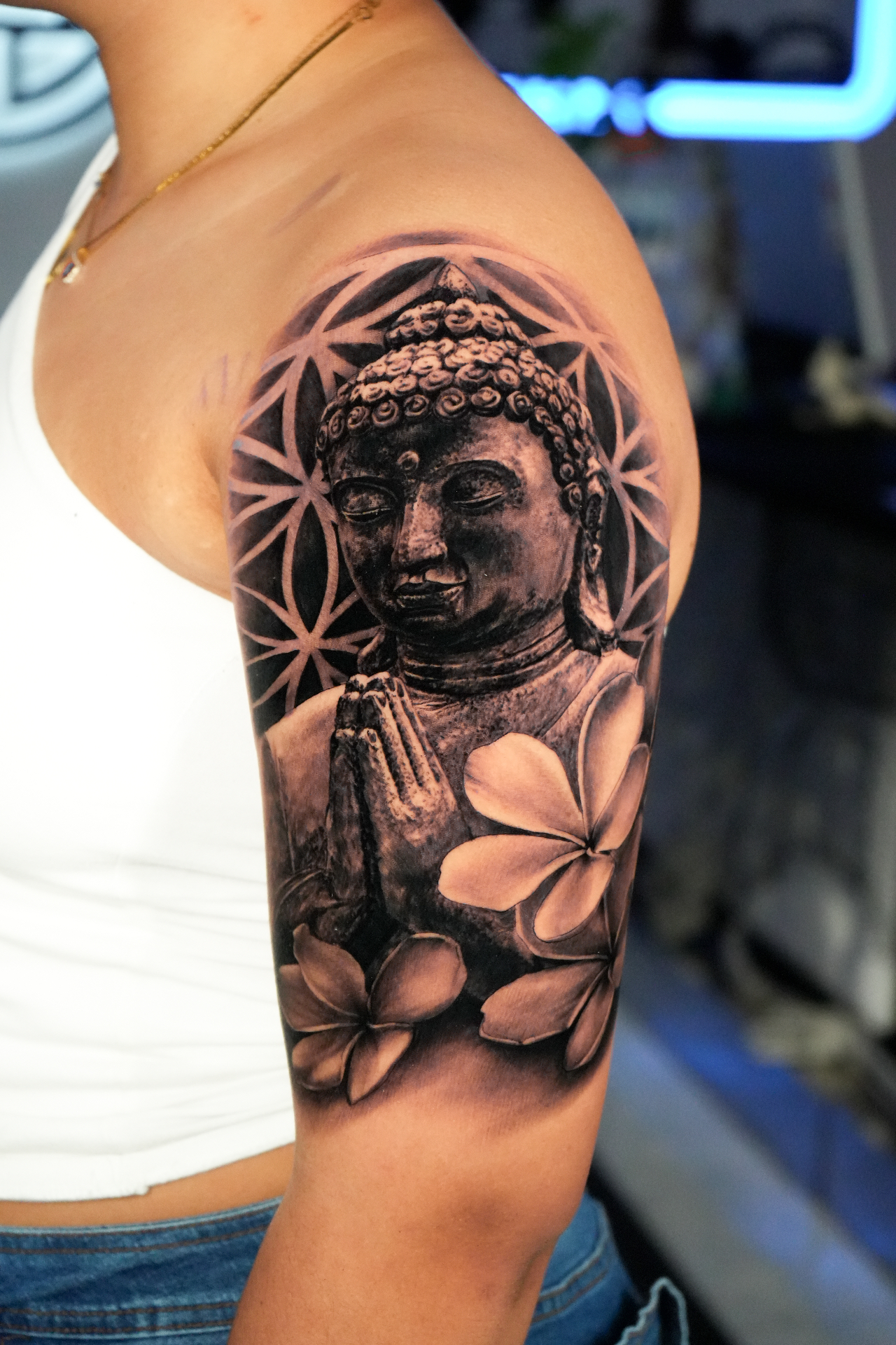 Black and grey realism upper arm piece of a stone-carved Buddha in prayer before sacred geometry, framed by plumeria blossoms